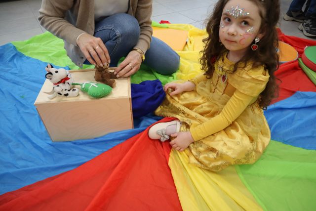 Na zdjęciu znajduje sie dziewczynka przebrana za księżniczkę siedząca na kolorowej chuście, obok niej klęczy kobieta i ustawia misia wraz z innymi małymi maskotkami na drewnianym pudełku.