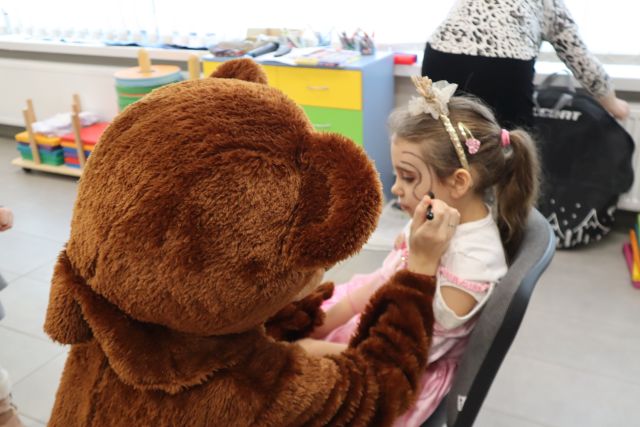 Na zdjęciu znajduje się osoba przebrana za misia, która maluje twarz dziewczynce przebranej za misia. W tle znajduje sie kobieta tyłem oraz szafka.