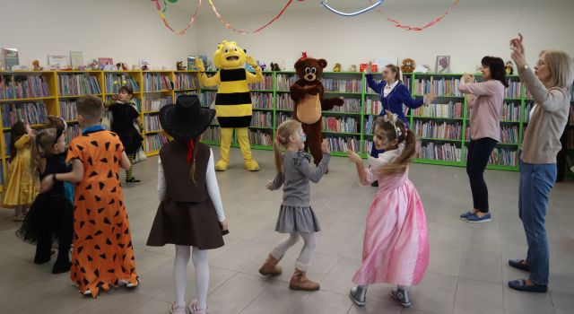 Na zdjęciu znajduje się grupka dzieci, trzy kobiety oraz miś z pszczółką i tańczą wspólnie kółku. W tle znajdują się półki z książkami.