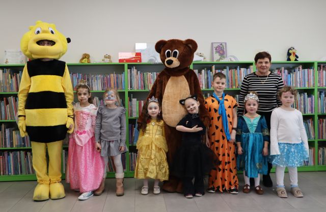 Na zdjęciu znajduje się siódemka dzieci w przebranich oraz kobieta i stoją do wspólnego zdjęcia z misiem i pszczółką. W tle znajduja się półki z książkami.
