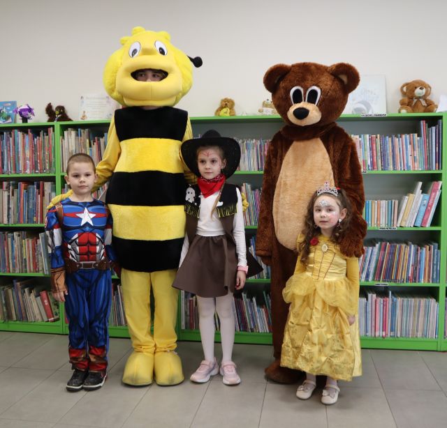 Na zdjęciu znajduje się trójka dzieci w przebranich stojąca do zdjęcia z misiem i pszczółką. W tle znajdują się półki z książkami.