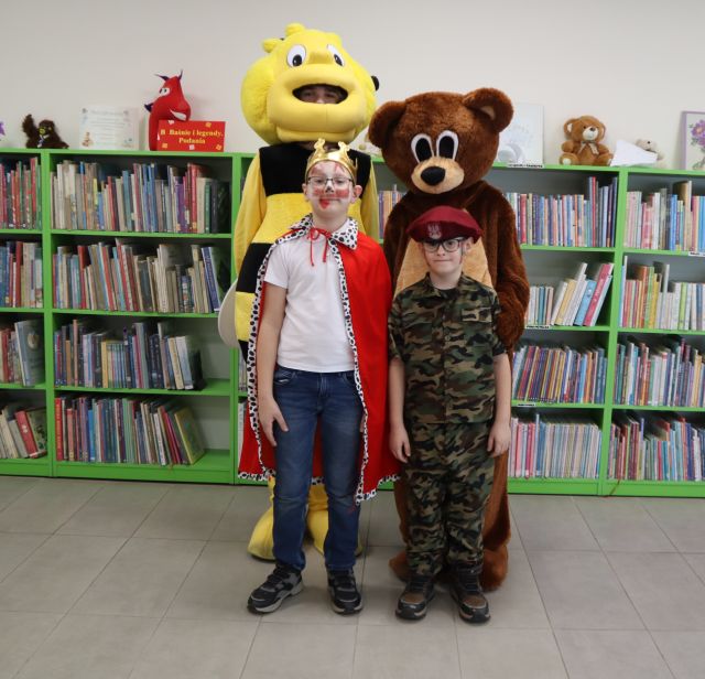 Na zdjęciu znajduje się dwójka chłopców, jeden przebrany za króla, a drugi za wojskowego i stoją do zdjęcia z misiem i pszczółką, którzy stoją za chłopcami. W tle znajdują się półki z książkami.