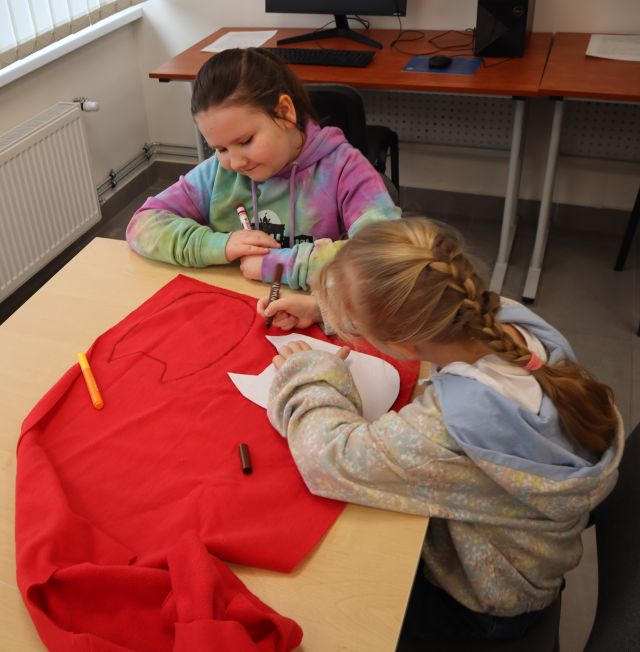 Na zdjęciu znajdują się dwie dziewcynki odrysowujące sowe na czerwonym materiale. Zajęcia odbywają się w sali komputerowej.