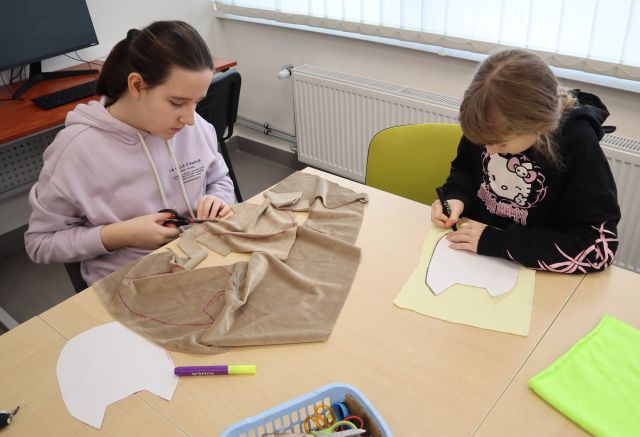 Na zdjęciu znajdują sie dwie dziewczynki przy stole, jedna z nich wycina sowe z materiału, a druga odrysowują ją na materiale. Warsztaty odbywają się w sali komputerowej.