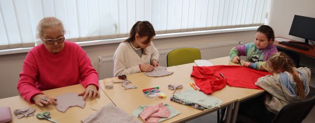 Na zdjęciu znajduje się trójka dziewczyn oraz jedna kobieta przy stole wycinając z materiałów dostępnych na stole sowy. Warsztaty odbywają się w sali komputerowej.