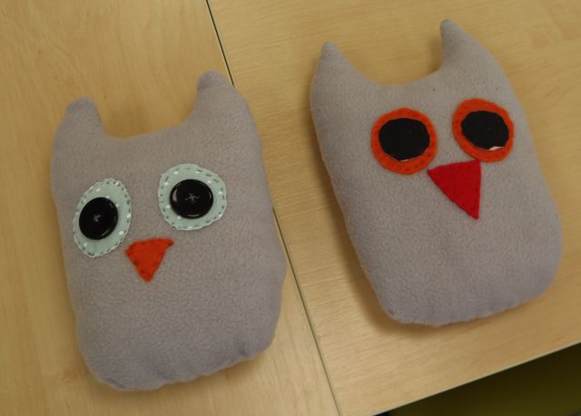 Na zdjęciu znajdują się dwie uszyte szare sowy z kolorowymi oczami i dziobem. Prace leżą na stole.