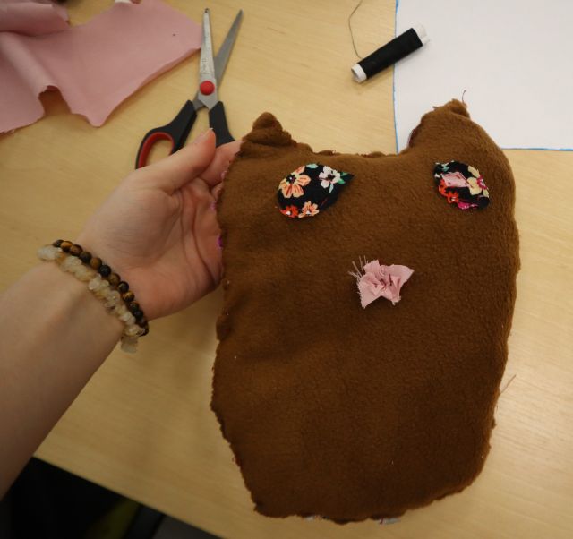 Na zdjęciu na tle stołu z materiałami i nożyczkami znajduje sie trzymana w ręcę uszyta brązowa sowa.