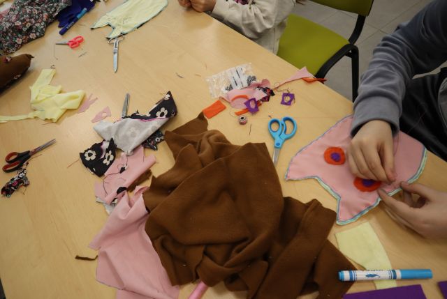 Na zdjęciu znajduje się stół wraz z kolorowymi materiałami, nożyczkami, przy stole siedzą dziewczynki, które szyją wycięte przez siebie z materiału sowy.