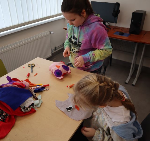 Na zdjęciu znajdują się dwie dziewczynki przy złączonych stołach, jedna z nich stoi i ma przed sobą dokończona uszytą sowe, a druga siedzi i kończy swoją prace. Na stole leżą resztki kolorowych materiałów, nożyczki oraz nici. Warsztaty odbywają się w sali komputerowej.
