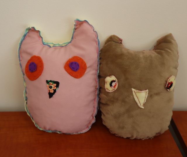 Na zdjeciu znajdują się dwie uszyte sowy obok siebie, jedna różówa a druga beżowa. Maskotki znajduja sie na drewnianym stole przy szarej ścianie.