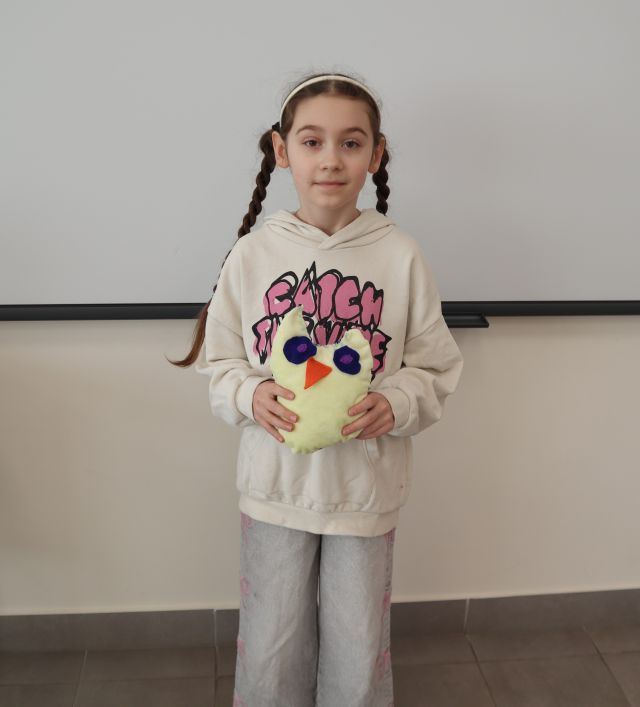 Na zdjęciu znajduje się dziewczynka stojąca i trzymająca w rękach uszytą przez siebie żółta maskotkę sowy. W tle znajduje się tablica interaktywna.