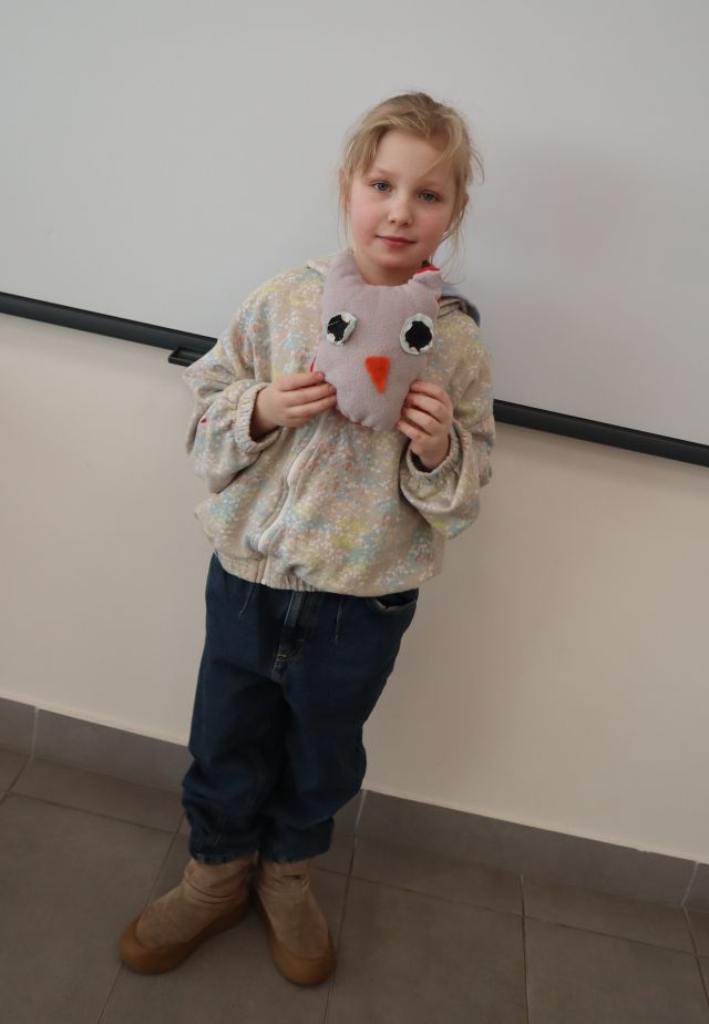 Na zdjęciu znajduje się dziewczynka stojąca i trzymająca w rękach uszytą przez siebie szarą maskotką sowy. W tle znajduje się tablica interaktywna. Zdjęcie zostało wykonane pod kątem.