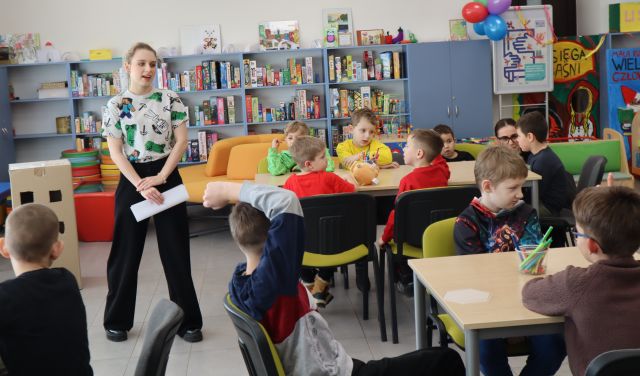 Na zdjęciu znajduje się grupa dzieci przy stolikach, po lewej stronie między stolikami stoi kobieta z kartką w ręce. W tle znajdują się półki z grami.