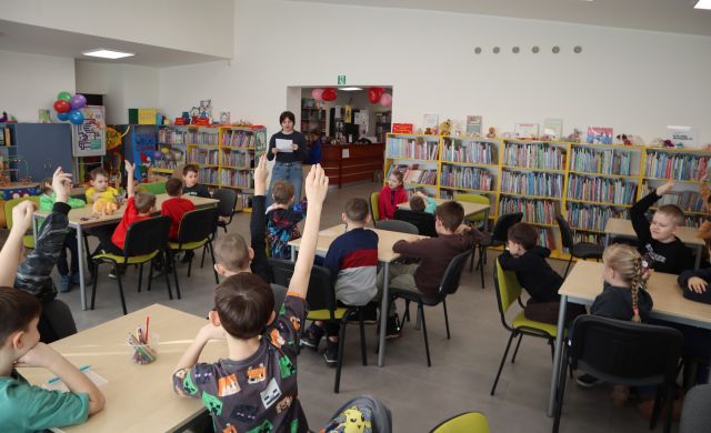 Na zdjęciu znajdują się dzieci przy sześciu stołach, część z nich ma podniesioną rękę, a w tle między stołami stoi kobieta i czyta pytania z kartki. W tle znajdują sie półki z książkami.