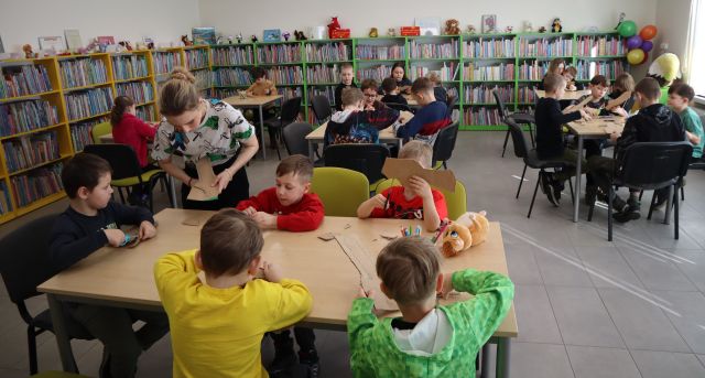 Na zdjęciu znajdują się dzieci przy stolikach, wycinające miescze z kartonu, przy pierwszym stoliku nachyla się kobieta i pomaga chłopcu w wycinaniu. W tle znajdują się półki z książkami.