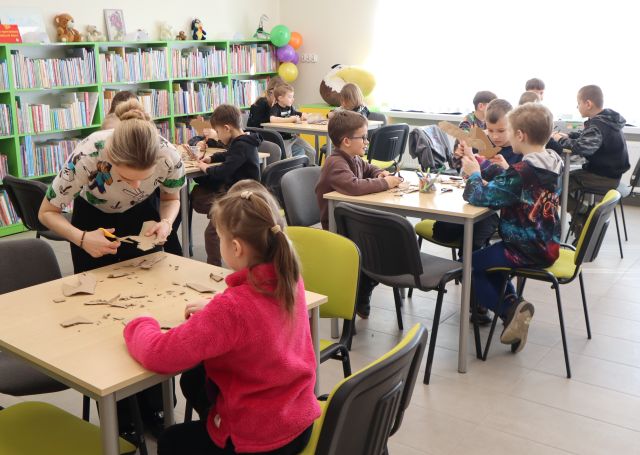 Na zdjęciu znajdują się dzieci przy pięciu stołach, uczestnicy wycinają miecze z kartonu. Przy pierwszym stoliku nachyla się kobieta i pomaga dziewczynce w wycinaniu. W tle znajdują się okna oraz półki z książkami.