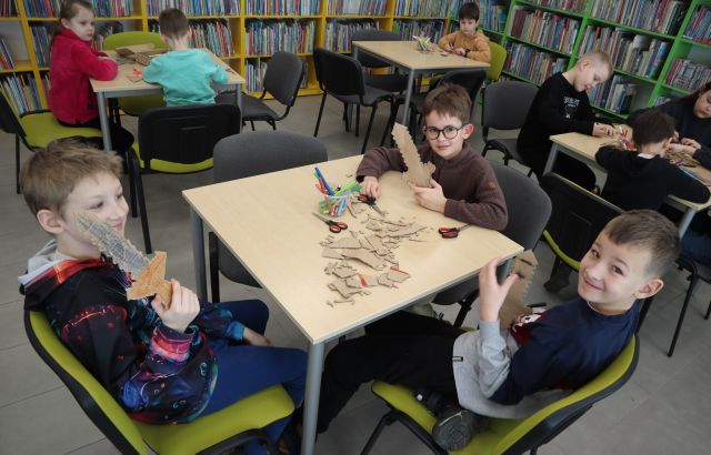 Na zdjęciu znajduje się trójka chłopców przy stoliku, którzy pokazują wykonane przez siebie miecze. Wokól nich znajdują sie stoliki z resztą uczestników, a w tle znajdują się półki z książkami.