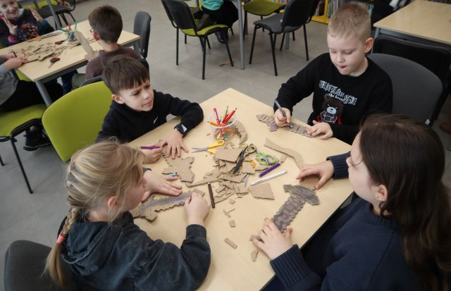 Na zdjęciu znajduje się stolik z czwórką dzieci, którzy kolorują wycięte przez siebie z kartonu miecze. W tle znajdują się stoły z resztą uczestników.