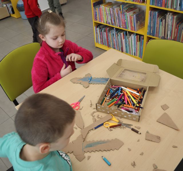 Na zdjęciu znajduje się dwójka dzieci przy stoliku i kolorują wycięte przez siebie z kartonu miecze. W tle znajdują się półki z książkami.