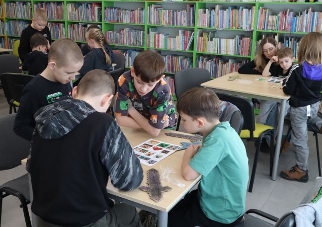 Na zdjęciu znajduja się dzieci przy stolikach, na kórych leżą wykonane przez nich miecze oraz kartki z kolorowymi obrazkami. W tle znajdują się półki z książkami.