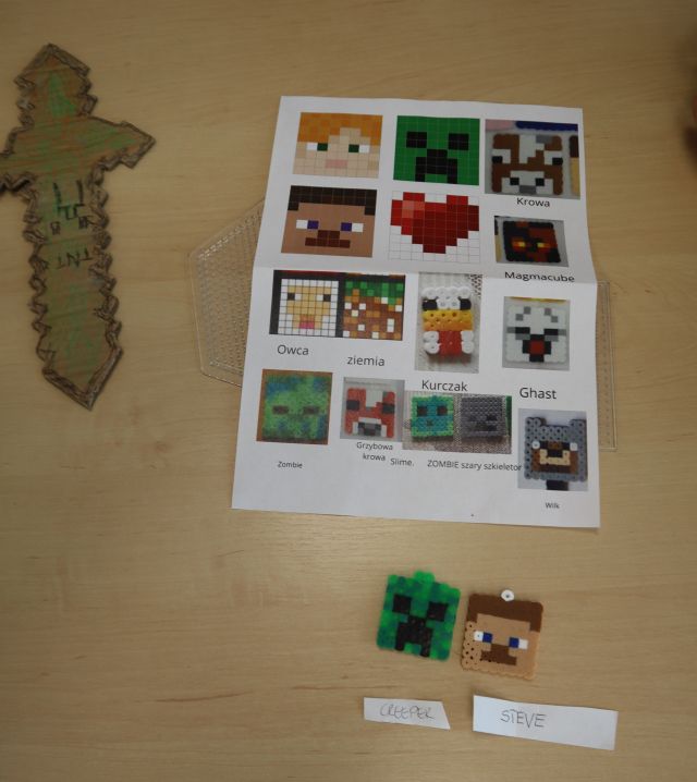 Na zdjęciu znajduje się leżąca na stole kartka z kolorowymi postaciami z Minecraft, pod nią leżą dwie postacie wykonane z koralików, a po lewej stronie leży pokolorowany kartonowy miecz.