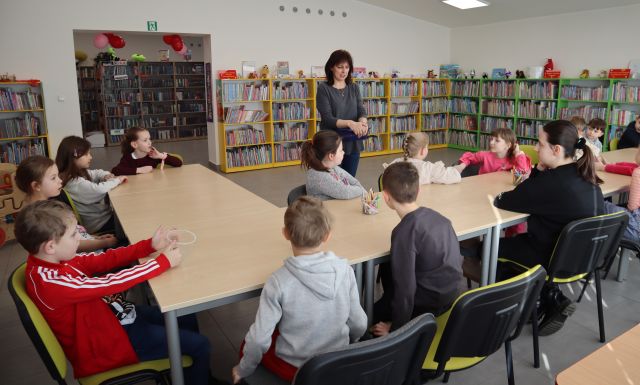 Na zdjęciu znajdują się dzieci przy stole ustawionym w literke L, przed stołem stoi kobieta, która trzyma w ręce mały niebieski koszyczek. W tle znajdują się półki z książkami.