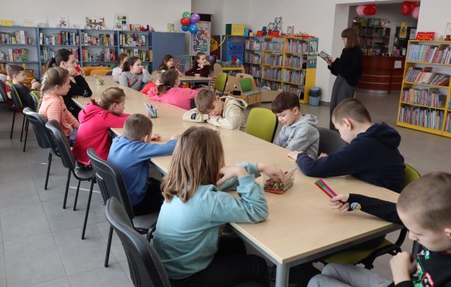 Na zdjęciu znajdują się dzieci przy złączonych w literkę L stolikach, przed nimi stoi kobieta, która czyta im fragment z trzymanej w rękach książki. W tle znajdują się półki z książkami.