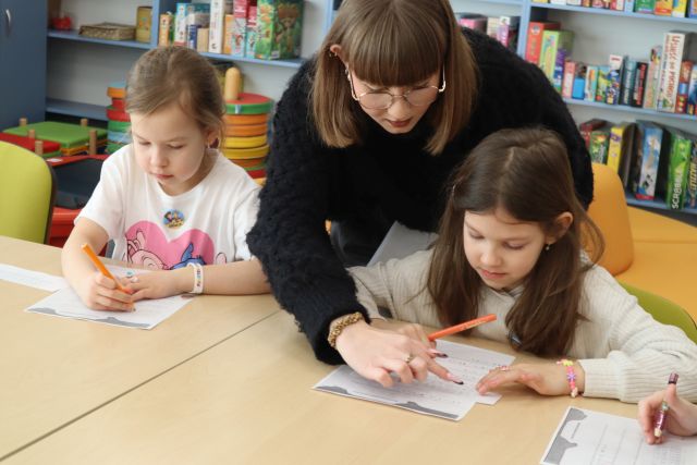 Na zdjęciu znajdują sie dwie dziewczynki przy złączonych stołach, przy jeden z nich nachyla się kobieta i wskazuje na kartkę, która leży na stole przed dziewczynką. W tle znajdują się półki z grami.