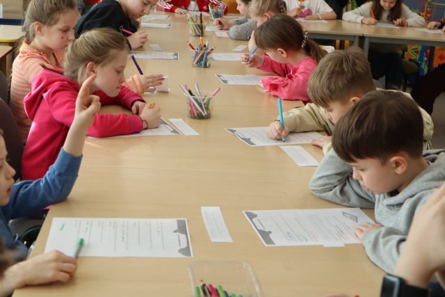 Na zdjęciu znajdują się dzieci przy złączonych stołach i rozwiązują zakodowane wiadomości ze znajdujących się przed nimi kartkach.
