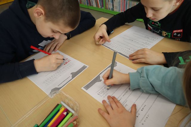Na zdjęciu znajduje sie trójka osób przy stoliku i rozkodowują zakodowane wiadomości z kartki, na stoliku znajduje się również pudełko z kredkami.