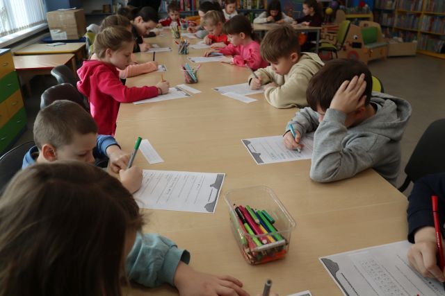 Na zdjęciu znajdują się dzieci przy złączonych stołach i rokodowują zakodowane wiadomości na kartkach, na stolikach znajdują się również kredki.