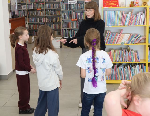Na zdjęciu znajdują się trzy dziewczynki stojące przodem do kobiety, która wskazuje na nie palcem. W tle znajdują się półki z książkami.