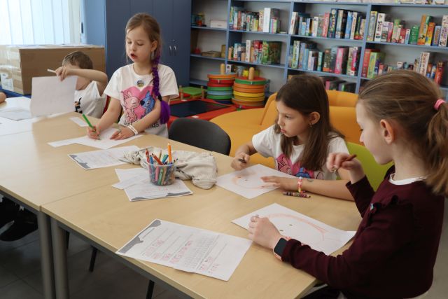 Na zdjęciu znajduje się czwórka dzieci przy złączonych stolikach i rysują one kobiete na kartkach, na stolikach znajduja się również kredki oraz kartki. W tle znajdują się półki z grami.