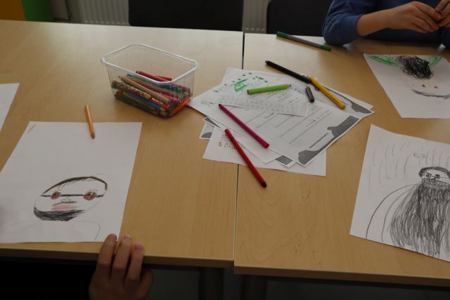 Na zdjęciu znajduje się złączone stoliki z rysunkami przedstawiającymi mężczyznę. Na środku stolika leży pudełko z kredkami oraz kartki z zakodowanymi wiadomościami.