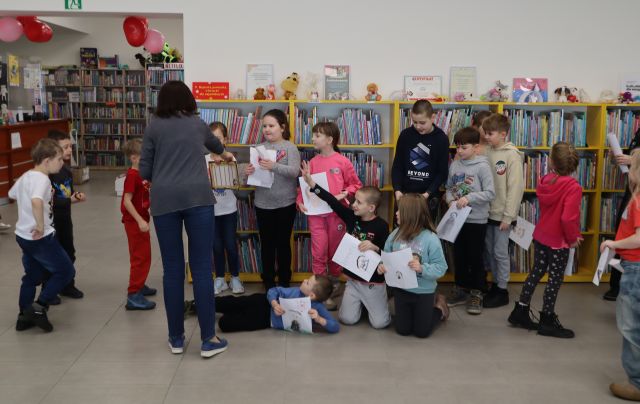 Na zdjęciu znajduje się grupa dzieci trzymające kartki oraz kobieta, która rozdaje im upominki. W tle znajdują się półki z książkami.