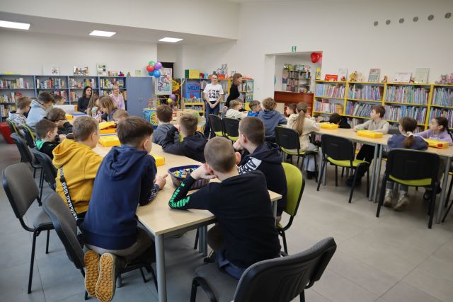 Na zdjęciu znajdują się dzieci siedzące przy złączonych w dwóch rzędach stołach, na których leżą klocki LEGO. W tle znajdują się dwie stojące kobiety oraz półki z książkami.