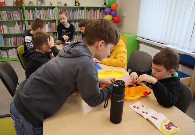 Na zdjęciu znajduje się grupka chłopców przy złączonych stolikach i układają LEGO. W tle znajdują się okna oraz półki z książkami.