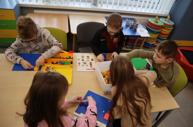 Na zdjęciu znajduje się piątka dzieci przy złączonych stolikach i wspólnie układają klocki LEGO. W tle znajdują się małe stoliki z kredkami.