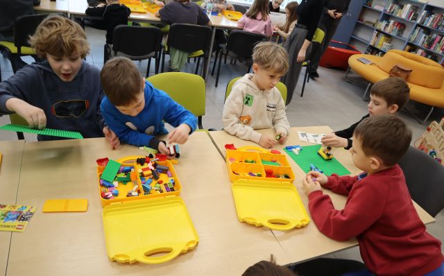 Na zdjęciu znajduje się piątka chłopców układających klocki LEGO przy złączonych stolikach. W tle znajduje się reszta uczetników przy stołach, a po prawej stronie okrągła żółta sofa, a za nią półki z grami.