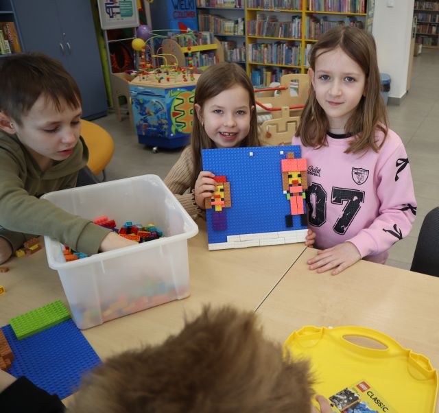 Na zdjęciu znajdują się dwie stojące przed złączonymi stołami dziewczynki, które pokazują wykonaną ilustracje z klocków LEGO przedstawiającą dwie rzucające się śnieżkami dziewczynki. Po lewej stronie znajduje się chłopiec sięgający klocek z pojemnika na stole. W tle znajdują się półki z ksiązkami oraz zabawki dla dzieci.