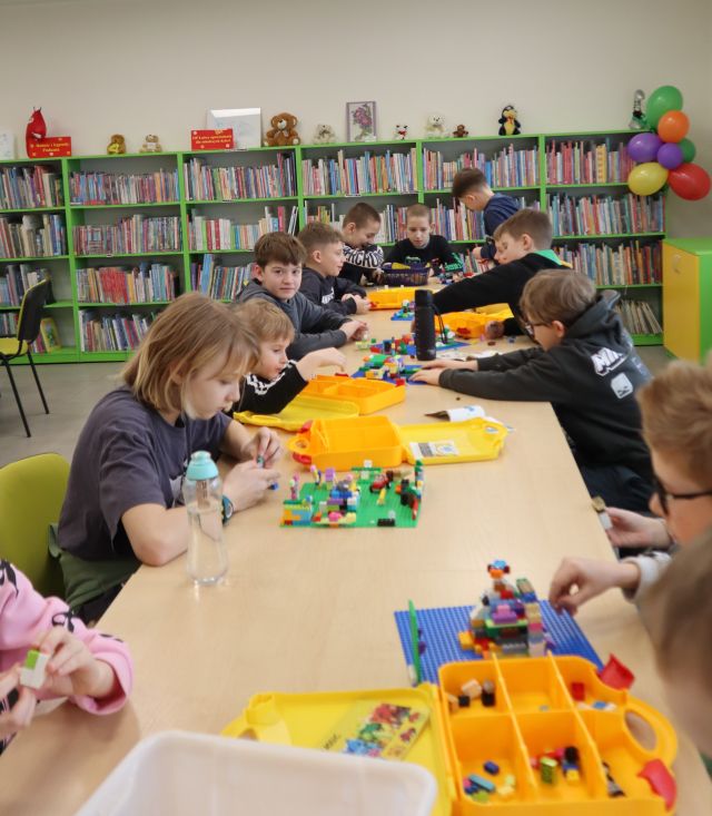 Na zdjęciu znajdują się dzieci siedzące przy złączonych stołach i układające LEGO. w tle znajdują się półki z książkami.