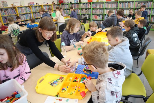 Na zdjęciu znajdują się dzieci siedzące przy złaczonych stolikach i układają LEGO, na przeciwko jednego chłopca nachyla sie kobieta z pojemniczkiem, z którego chłopiec losuje kartkę. W tle znajdują się półki z książkami oraz drugi rząd stołów z uczestnikami.