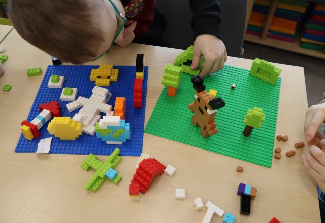 Na zdjęciu znajdują się dwie budowle z LEGO, jedna przedstawia kosmos, druga las, dookoła na stole leżą klocki LEGO, a nad pracami nachyla się chłopiec.