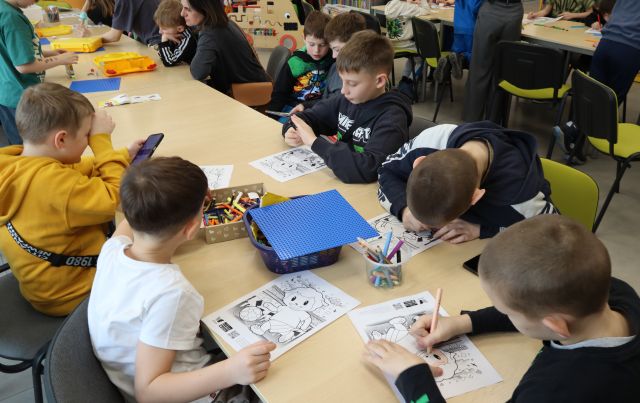 Na zdjęciu znajdują się dzieci przy złączonych stołach, część z nich koloruje kolorowanki, a część układa klocki LEGO.