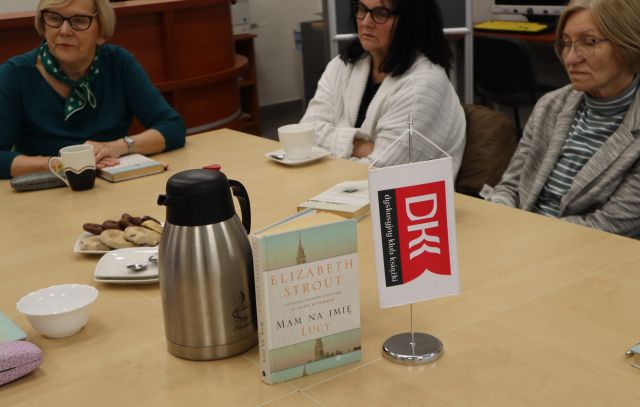 Na zdjęciu znajduje się stojący na stole proporczyk z Dyskusyjnego Klubu Książki, książka Mam na imię Lucy oraz termosy i szklanki, przy złączonych stołach siedzą trzy kobiety.