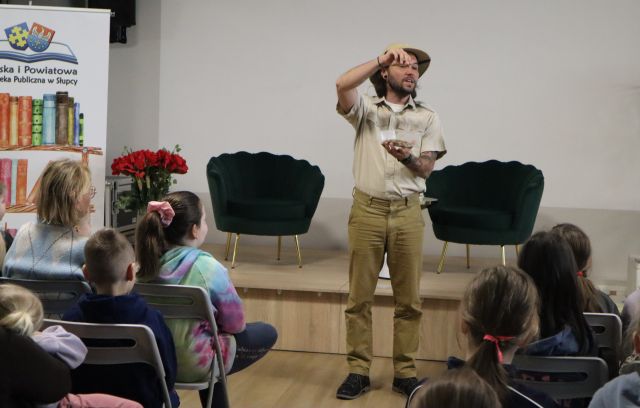 Na zdjęciu znajduje się mężczyzna, który trzyma w ręku pudełko z robakami, przed nim w dwóch rzędach stoją krzesła z uczestnikami, a w tle na małej scenie stoją fotele, a po lewej stronie obok sceny baner z książkami i logiem biblioteki.