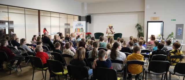 Na zdjęciu znajduje się grupa dzieci i kilka dorosłych, którzy znajdują się na krzesłach na holu biblioteki, przed nimi stoi mężczyzna.