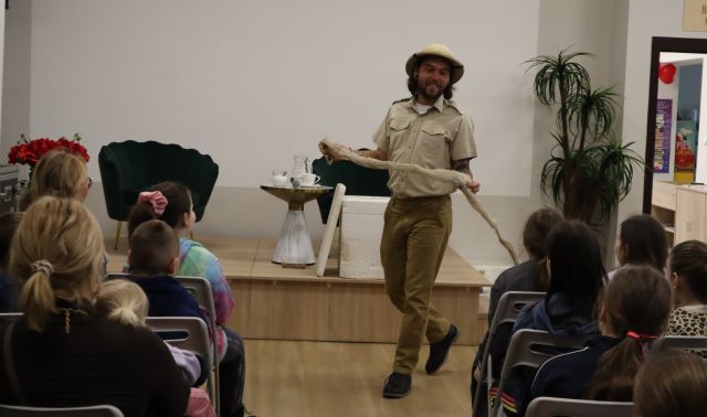 Na zdjęciu znajduje się mężczyzna w ruchu, który trzyma w rękach wylinkę węża. Przed nim w dwóch rzędach siedzą dzieci na krzesłach. W tle za mężczyzną znajduje się mała scena z fotelami.