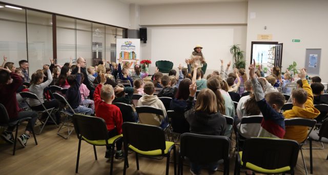 Na zdjęciu znajduje się grupa dzieci na krzesłach, część z nich ma podniesioną rękę, a przed nimi stoi mężczyzna z wężem. Spotkanie odbywa się na holu biblioteki.