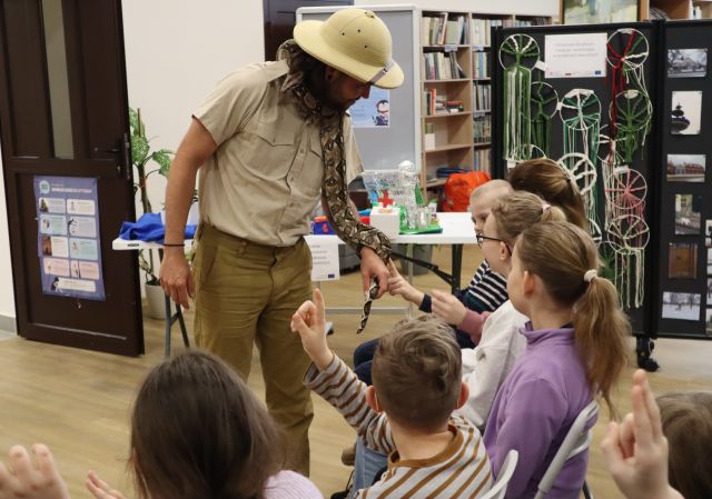 Na zdjęciach znajduje się mężczyzna z wężem przed dziećmi, które głaszczą węża. W tle znajduje się makieta miasta oraz wystawa drzew na kole z makramy i zdjęcia.